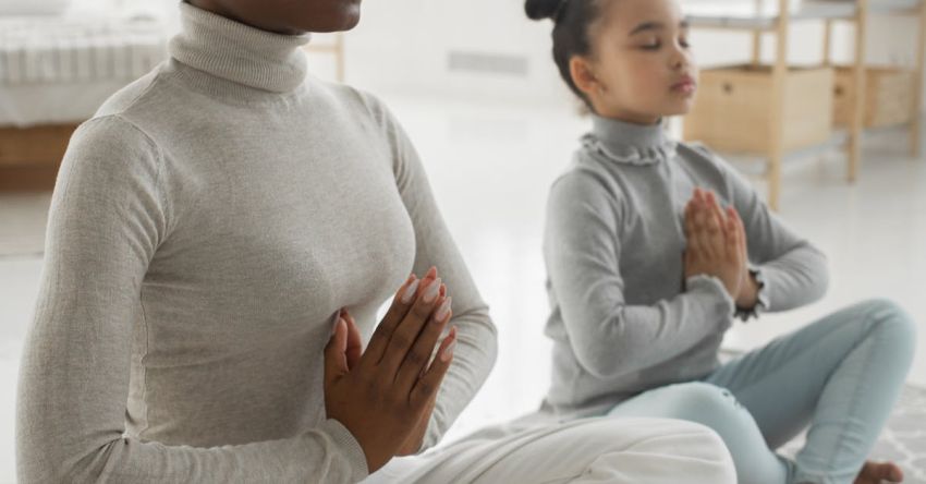 Kids Mindfulness - Full body of tranquil young barefooted African American woman in casual clothes sitting on carpet in Padmasana pose with closed eyes while meditating with adorable little daughter at home